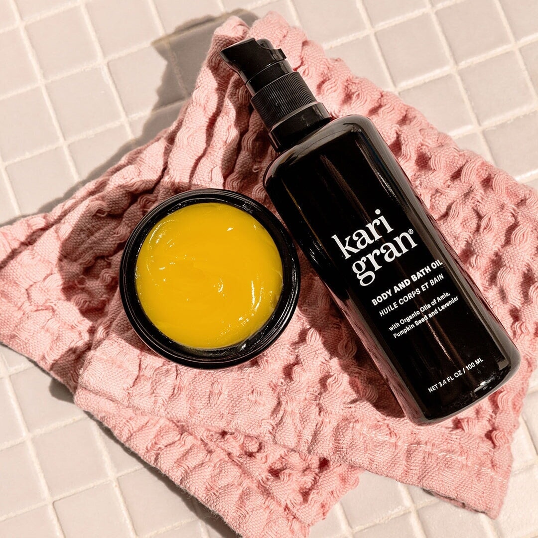 kari gran body and bath oil next to the essential serum balm on a pink washcloth