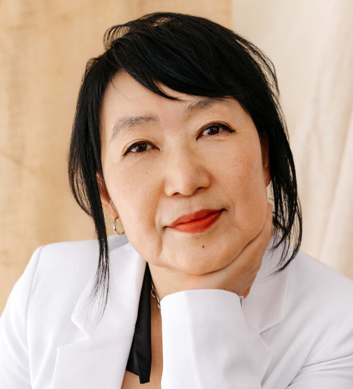 Woman with black hair and red lipstick wearing a white blazer against a beige background